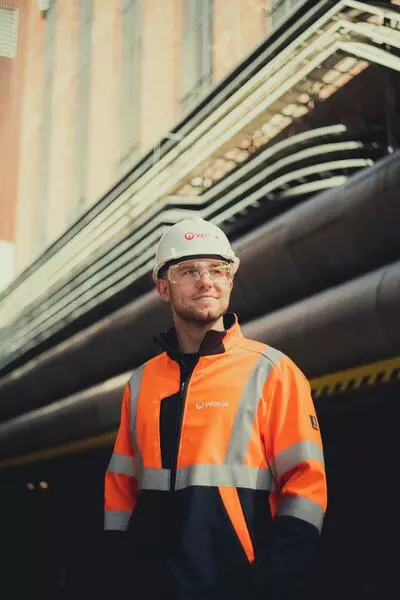 Technicien Veolia Énergie Performance en EPI complets — casque blanc, lunettes de sécurité et veste haute visibilité orange et marine — sur site industriel, devant un réseau de canalisations.